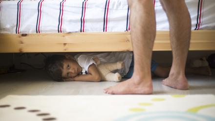 Ein Kind versteckt sich unter einem Bett. Vor dem Bett sind die nackten Beine und Füße eines Mannes zu sehen. Das Kind sieht ängstlich aus.