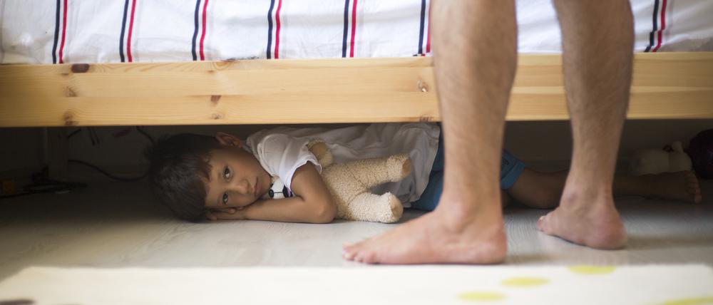 Ein Kind versteckt sich unter einem Bett. Vor dem Bett sind die nackten Beine und Füße eines Mannes zu sehen. Das Kind sieht ängstlich aus.