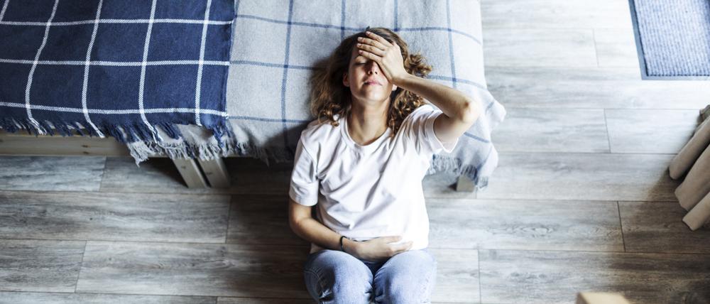 Eine junge Frau sitzt erschöpft auf dem Fußboden neben einem Bett (Symbolbild).