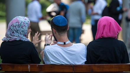 Zwei junge Muslimas mit Kopftuch und ein junger jüdischer Mann mit Kippa unterhalten sich miteinander.