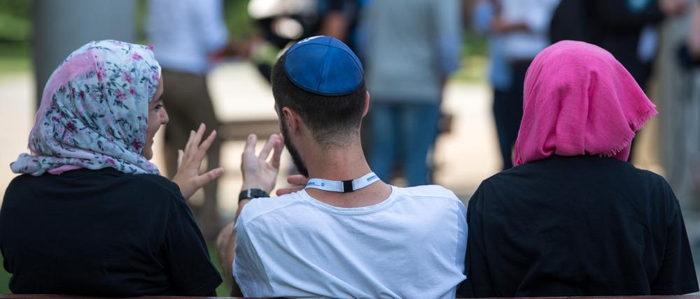 Zwei junge Muslimas mit Kopftuch und ein junger jüdischer Mann mit Kippa unterhalten sich miteinander. 
