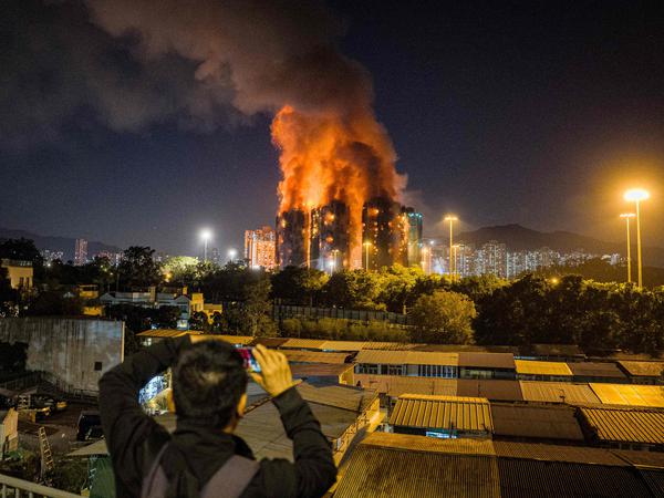 An onlooker takes photos as a major fire engulfs several apartment blocks at the Wang Fuk Court residential estate (background) in Hong Kong's Tai Po district on November 26, 2025. At least four people were killed when a fire engulfed several high-rise blocks in a Hong Kong residential estate on November 26, the government said, with media reporting that some residents were trapped inside. (Photo by AFP)