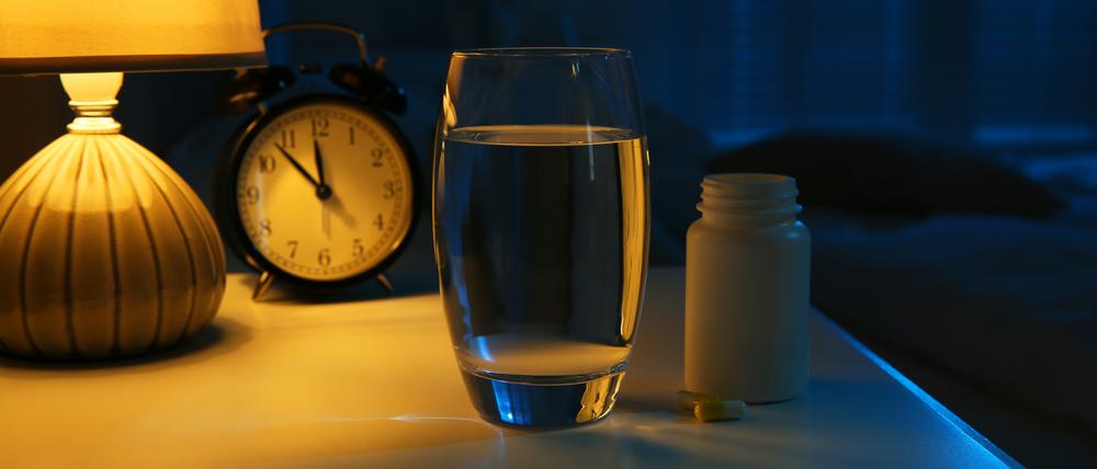 Illustration: Eine Tablettenbox, ein Glas Wasser und ein Wecker stehen auf einem Nachttisch.