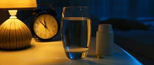 Illustration: Eine Tablettenbox, ein Glas Wasser und ein Wecker stehen auf einem Nachttisch.