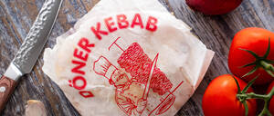 Eine Dönerverpackung liegt neben einem Küchenmesser, Tomaten, Knoblauch und einer Zwiebel.