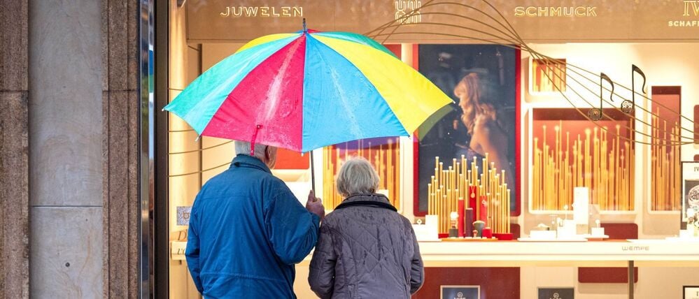 Zwei ältere Personen stehen bei Regen vor einem Schaufenster eines Schmuckgeschäfts und betrachten die ausgestellten Waren (Symbolbild).