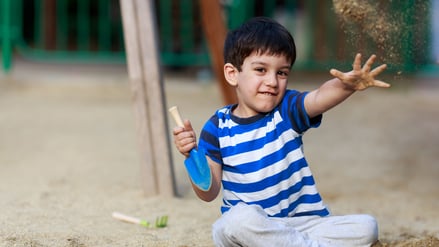 Ein etwa fünfjähriger Junge sitzt auf einem Spielplatz und wirft mit Sand. Er trägt ein blau gestreiftes T-Shirt.
