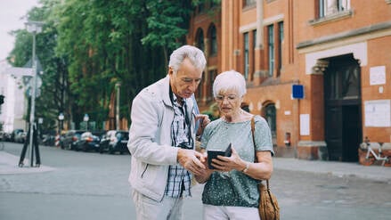 Ein älteres Ehepaar steht auf einer Straße und blickt suchend auf ein Smartphone.