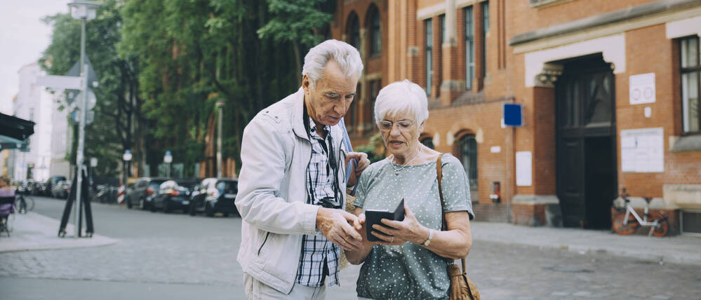 Ein älteres Ehepaar steht auf einer Straße und blickt suchend auf ein Smartphone.
