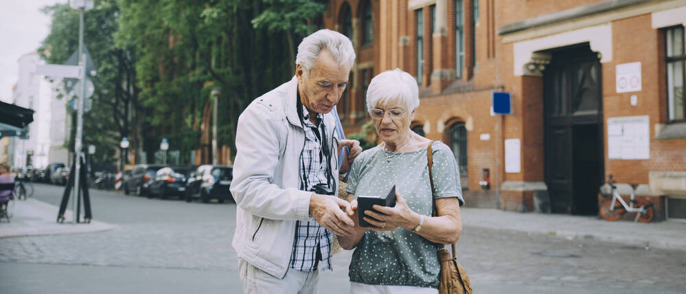 Ein älteres Ehepaar steht auf einer Straße und blickt suchend auf ein Smartphone.