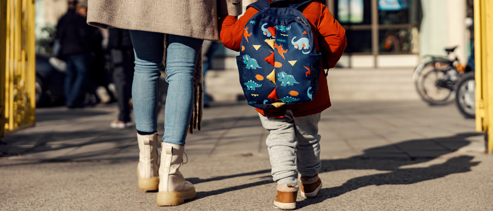 Ein Kind mit Rucksack geht an der Hand seiner Mutter auf den Eingang einer Kita zu.