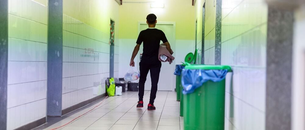Ein Migrant geht in der Zentralen Aufnahmeeinrichtung für Asylbewerber in Bayern mit der Erstausstattung an Besteck, Bettwäsche und Hygieneartikeln über einen Gang zu seinem Wohnbereich (Symbolbild).