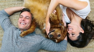 Ein fröhliches Paar liegt auf dem Teppich und kuschelt mit einem Hund, der zwischen ihnen liegt.