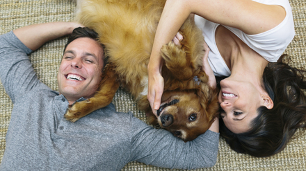 Ein fröhliches Paar liegt auf dem Teppich und kuschelt mit einem Hund, der zwischen ihnen liegt.