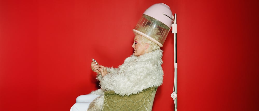Senior woman sitting under hairdryer using mobile phone, side view