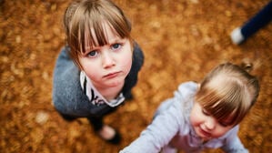 Zwei Mädchen im Kitaalter schauen den Betrachter unzufrieden an. Im Hintergrund ist braunes Laub zu sehen.