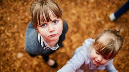 Zwei Mädchen im Kitaalter schauen den Betrachter unzufrieden an. Im Hintergrund ist braunes Laub zu sehen.