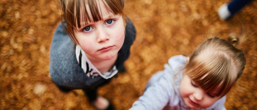 Zwei Mädchen im Kitaalter schauen den Betrachter unzufrieden an. Im Hintergrund ist braunes Laub zu sehen.