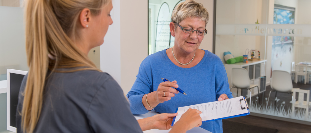 Eine Patientin reicht einer Arzthelferin einen Anamnesebogen.
