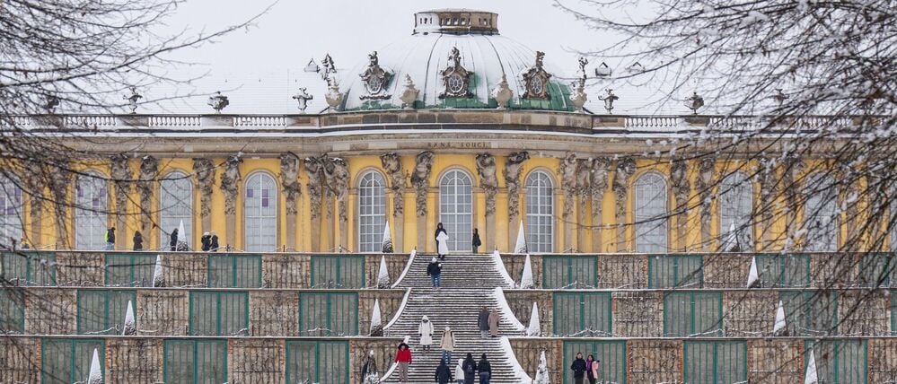 Menschen gehen vor der verschneiten Kulisse des Schloss Sanssouci in Potsdam spazieren.
