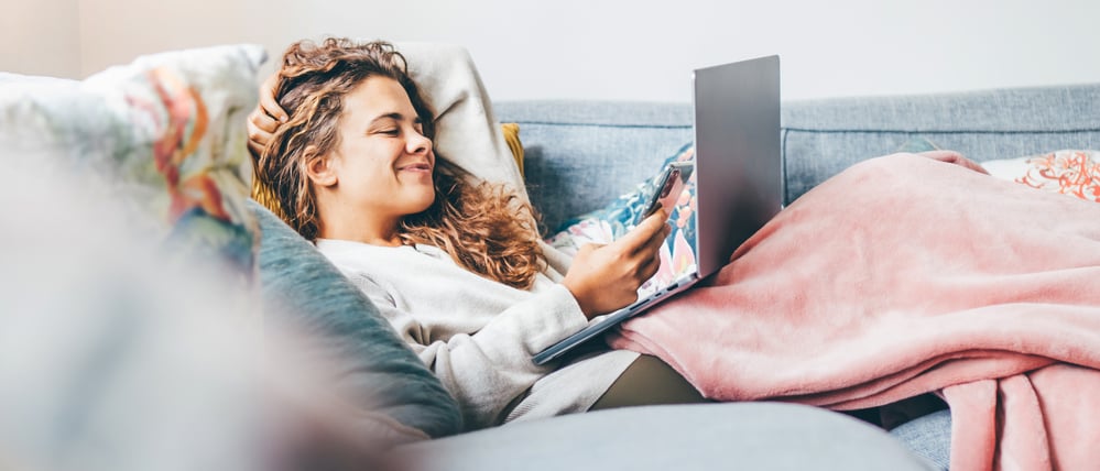 Woman wrapped in blanket using laptop on sofa at home.