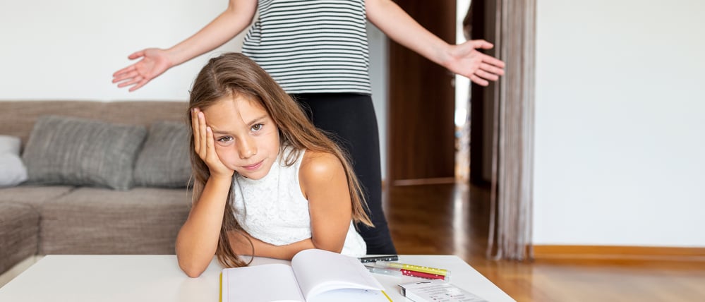 Ein etwa zehnjähriges Mädchen sitzt gestresst vor ihren Unterlagen am Wohnzimmertisch. Hinter ihr breitet ihre Mutter resigniert die Arme aus.