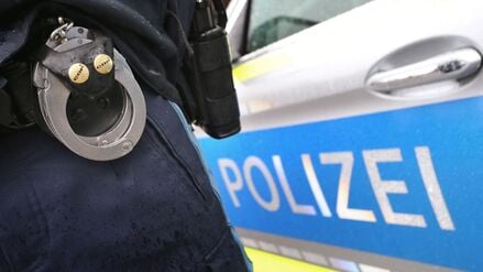 Ein Polizist mit Handschellen und einer Pistole am Gürtel steht vor einem Streifenwagen (Symbolbild).
