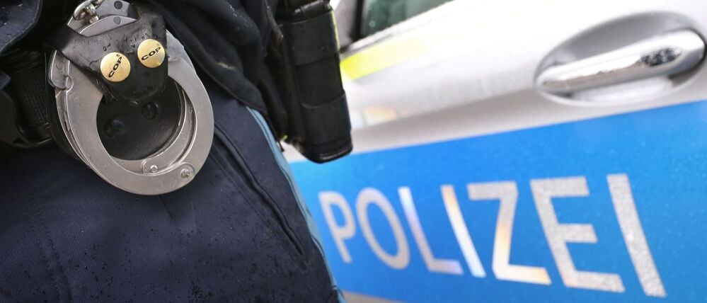 Ein Polizist mit Handschellen und einer Pistole am Gürtel steht vor einem Streifenwagen (Symbolbild).