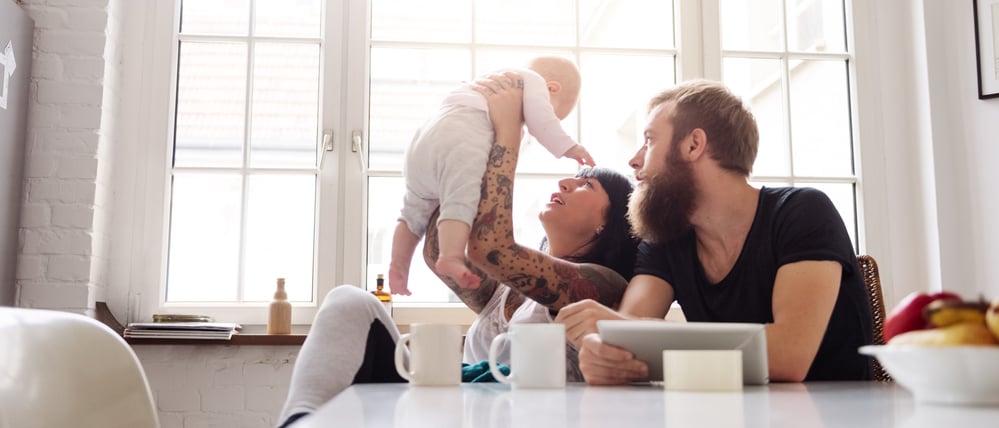 Ein junges Paar mit Baby sitzt am Küchentisch. Die Mutter hebt das Baby in die Höhe und schaut es an, der Vater schaut skeptisch zu.
