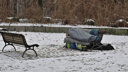 Eine obdachlose Person liegt in einem Schlafsack auf einer Parkbank. (Symbolbild)