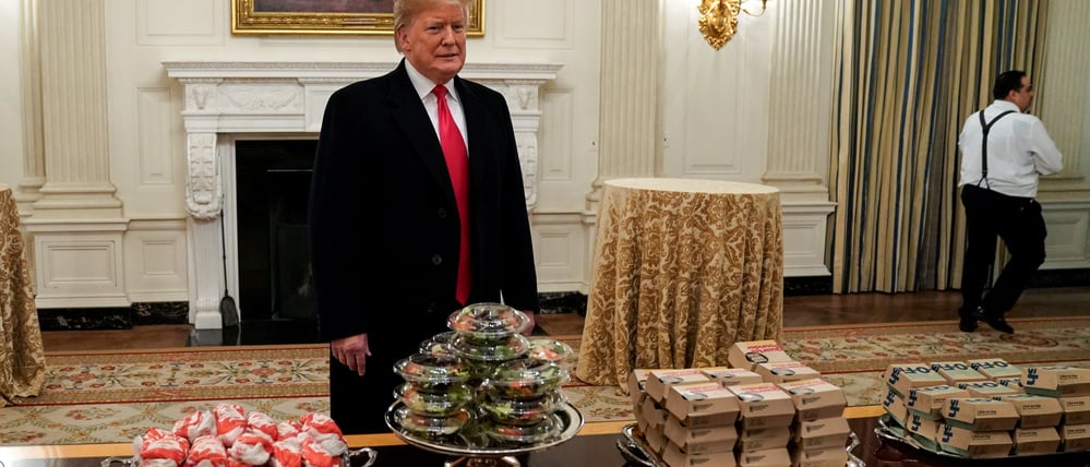 US-Präsident Donald Trump spricht hinter Stapeln von McDonald’s‑Hamburgern, Chick‑fil‑A‑Sandwiches und anderem Fast Food, als er im Jahr 2019 Mitglieder des Footballteams der North Dakota State University im Weißen Haus empfängt.