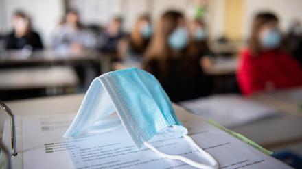 ARCHIV - 17.11.2020, Bayern, München: ILLUSTRATION - Eine Maske liegt im Unterricht auf Unterlagen, während im Hintergrund Schülerinnen und Schüler in einem Gymnasium mit Mund- und Nasenschutz zu sehen sind. (zu dpa «Große Mehrheit der Schüler sieht Lernrückstände durch Schließungen») Foto: Matthias Balk/dpa +++ dpa-Bildfunk +++