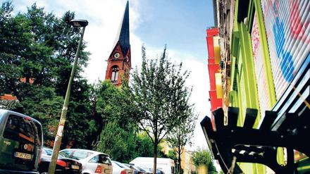 Immanuelkirchstraße in Prenzlauer Berg: Einfallstor und Trutzburg. Blick auf die Immanuelkirche und den oberen Abschnitt der Immanuelkirchstraße (Richtung Prenzlauer Allee). Rechts erkennt man die roten Erker des Immanuelkirch-Carrés. Dieser 2008 fertiggestellte Komplex ist Inbegriff für die Gentrifizierung der Straße.