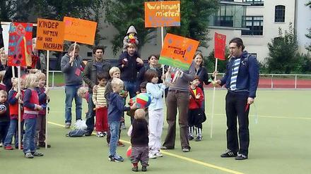 Mit Plakaten und Trillerpfeifen demonstrieren Eltern und Kinder auf dem Platz an der Körtestraße, im Hintergrund die Wohnhäuser.