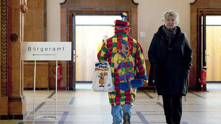 In einem Gebäude: Ein Bürgeramtschild, daneben ein Clown und eine Frau, die nicht lacht. 