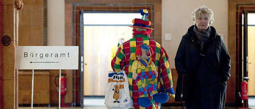 In einem Gebäude: Ein Bürgeramtschild, daneben ein Clown und eine Frau, die nicht lacht. 
