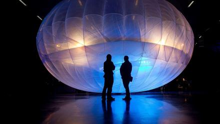 Zwei Personen, die nur als Schatten zu erkennen sind, stehen in einem dunklen Raum vor einem farbig erleuchteten Ballon. Der Ballon hat die Form einer Ellipse und überragt die Personen bei weitem.