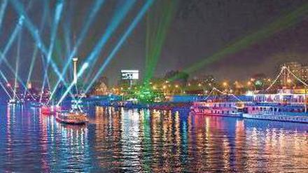 Bunte Schiffe auf dem glitzernden Wasser, bunte Scheinwerfer, die in den nächtlichen Himmel strahlen, hinten sieht man den Fernsehturm.