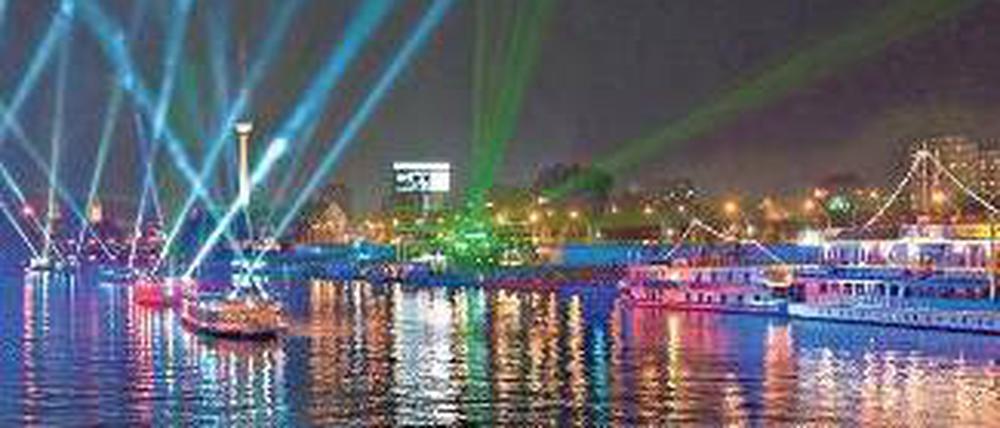 Bunte Schiffe auf dem glitzernden Wasser, bunte Scheinwerfer, die in den nächtlichen Himmel strahlen, hinten sieht man den Fernsehturm.