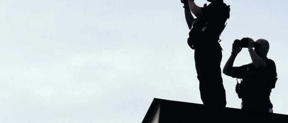 Zwei bewaffnete Männer mit riesigen Ferngläsern stehen im Gegenlicht auf einem Dach.