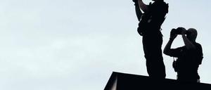 Zwei bewaffnete Männer mit riesigen Ferngläsern stehen im Gegenlicht auf einem Dach.
