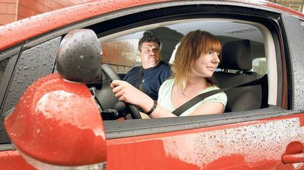 Eine junge Frau sitzt in einem roten Auto und schaut über ihre Schulter, neben ihr ein älterer Mann. 