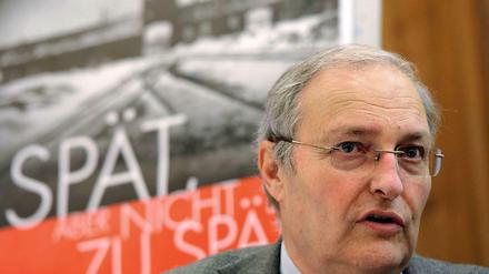 Efraim Zuroff, Direktor des Wiesenthal-Zentrums in Israel, vor dem Plakat der Kampagne. Auf dem Plakat steht: "Spät, aber nicht zu spät", im Hintergrund das Tor des Vernichtungslagers Auschwitz. 