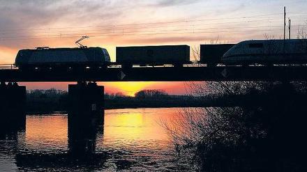 Ein Containerzug fährt bei Sonnenuntergang über eine Aller-Brücke und wird von einem ICE überholt.