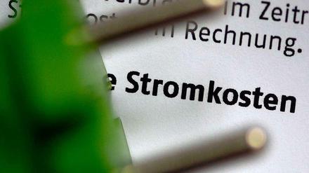 Ein grüner Stecker vor einem Papier, auf dem das Wort "Stromkosten" zu lesen ist. 
