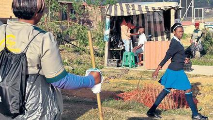 In einem Township in Johannesburg steht eine Gartenarbeiterin an einem Weg, auf dem ein Mädchen in Schuluniform vorbeigeht.