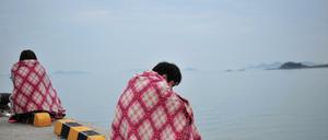 Überlebende des Fährunglücks sitzen nach ihrer Rettung auf einer Hafenmole und schauen auf das Meer hinaus.