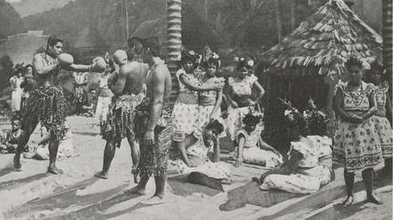 Eine Gruppe von Männern und Frauen aus Samoa stehen und sitzen in einer Südsee-Kulisse, zwei Männer tragen Boxhandschuhe und nehmen eine Kampfpose ein.