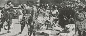 Eine Gruppe von Männern und Frauen aus Samoa stehen und sitzen in einer Südsee-Kulisse, zwei Männer tragen Boxhandschuhe und nehmen eine Kampfpose ein.