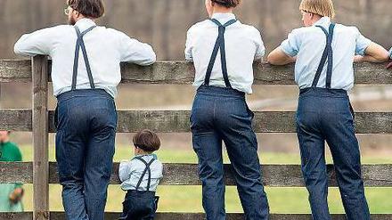 Drei erwachsene Amish und ein Kind stehen mit dem Rücken zum Betrachter an einem Zaun.